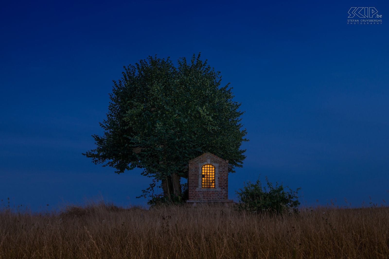 Hageland by night - Saint Joseph chapel Under an old lime tree in the middle of the fields in the village of Sint-Pieters-Rode (Holsbeek) the Saint Joseph's chapel is located. The simple chapel consists of bricks and a sandstone frame. It dates from the early 19th century. This photo was made July in the beginning of the blue hour (the hour after sunset). Stefan Cruysberghs